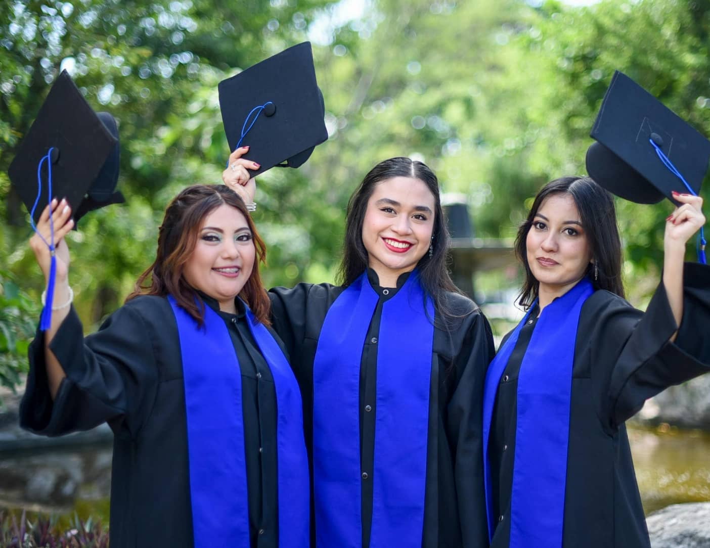 Three female graduates in blue academic gowns and caps celebrating by holding their caps up in the air.