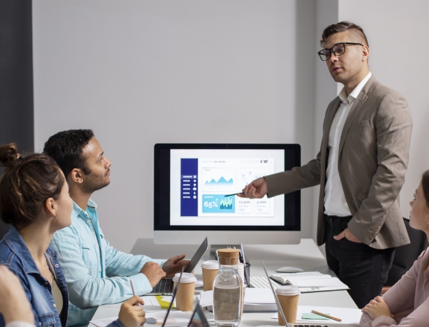 A professional instructor presenting business data and charts to a group of colleagues in a modern office meeting.