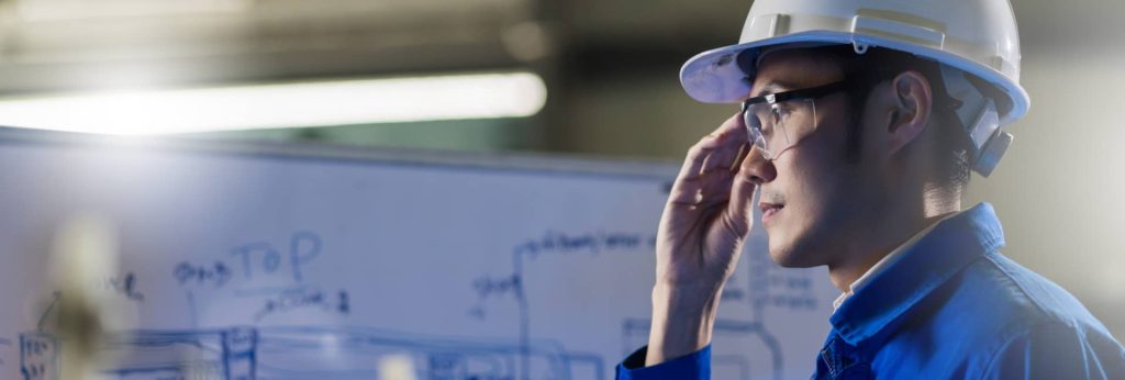 A civil engineer wearing a hard hat and safety glasses reviewing construction design blueprints on a whiteboard.