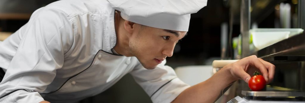 A young male chef in a white uniform and hat carefully prepping ingredients at a stainless steel station in a commercial kitchen.