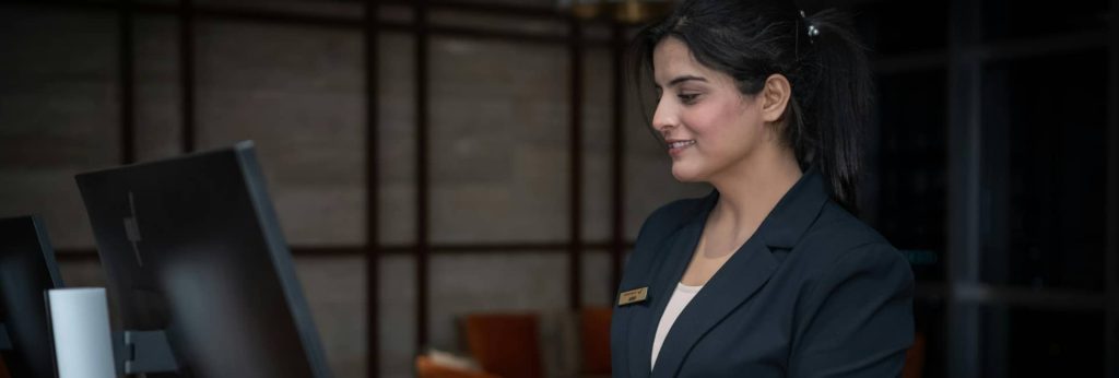 A professional female hotel receptionist smiling at the front desk service counter during hospitality operations training.