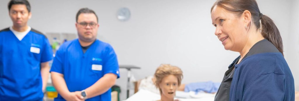Nursing students in blue scrubs practicing patient care on a medical manikin with an instructor during a practical training lab.