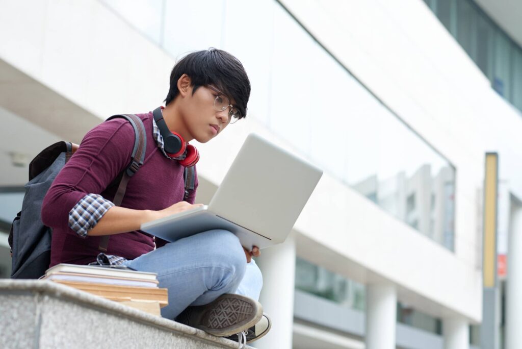 A male university student with headphones sitting on outdoor campus steps and working on his laptop.
